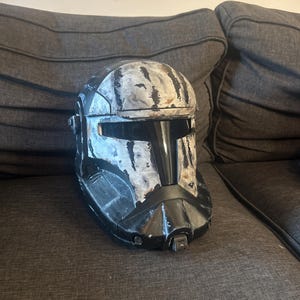 May include: A black and grey helmet with a distressed, textured appearance. The helmet features a visor and is positioned on a grey surface. The helmet seems to be a replica or prop.