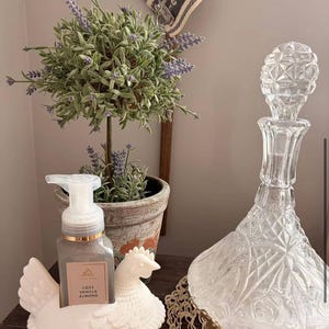 May include: A decorative arrangement featuring a clear glass decanter with a diamond pattern, a white ceramic hen-shaped soap holder with a soap dispenser, and a small potted plant with lavender-colored flowers.