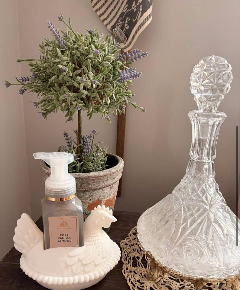 May include: A decorative arrangement featuring a clear glass decanter with a diamond pattern, a white ceramic hen-shaped soap holder with a soap dispenser, and a small potted plant with lavender-colored flowers.