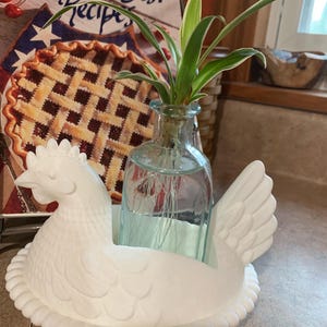 May include: A white ceramic hen-shaped planter holding a small glass bottle with a green plant. The hen is sitting on a white base. A cookbook titled "America's Best Recipes" is in the background.