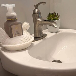 May include: A white ceramic hen soap dish holds a brown soap dispenser with a white pump. The soap dispenser has a label that reads "Cozy Vanilla Almond." A silver faucet and a small potted succulent are also visible.