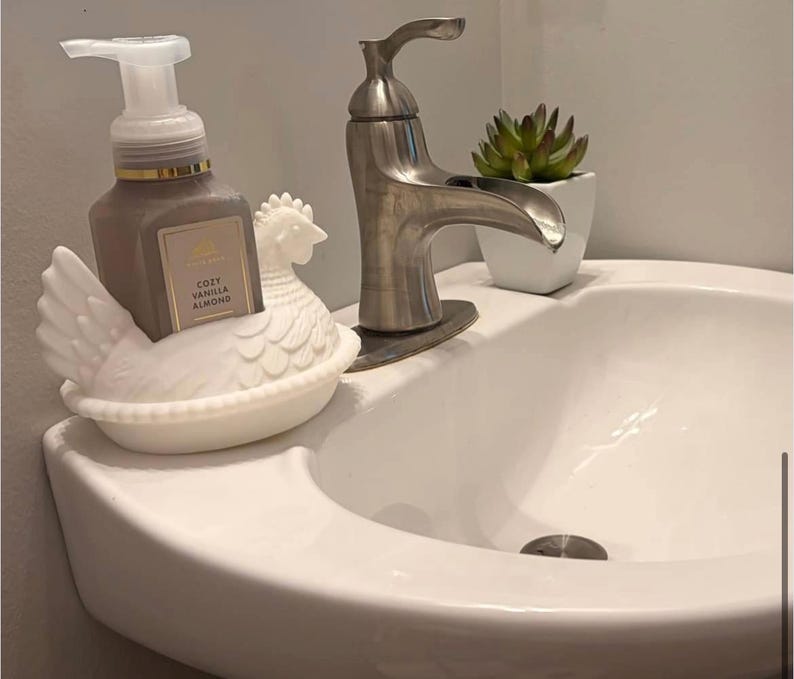 May include: A white ceramic hen soap dish holds a brown soap dispenser with a white pump. The soap dispenser has a label that reads "Cozy Vanilla Almond." A silver faucet and a small potted succulent are also visible.