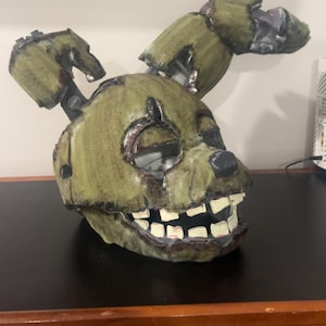 May include: A weathered olive green rabbit mask featuring a distressed texture, large hollow eye sockets, a small black nose, and a wide grin with square white teeth. The mask includes two tall, segmented, and partially broken rabbit ears.