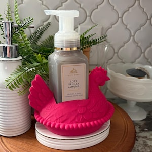 May include: A gray foaming hand soap bottle with a gold rim and white pump sits in a bright pink chicken-shaped holder. The soap is labeled "Cozy Vanilla Almond." A white ceramic soap dispenser and greenery are in the background.