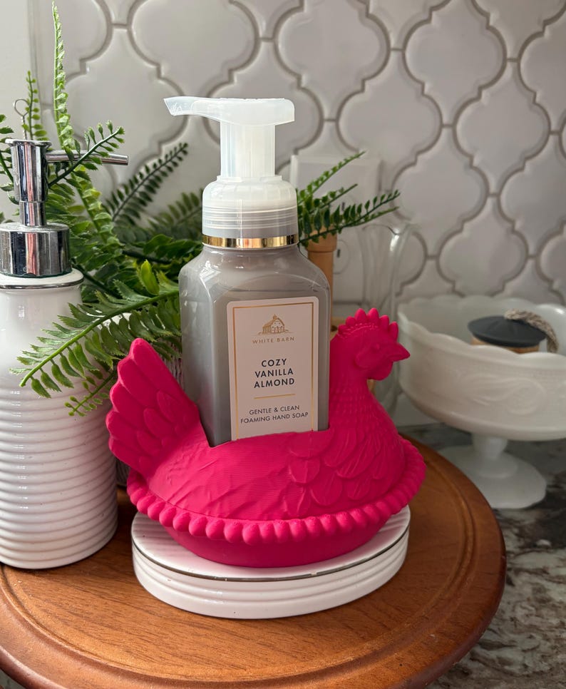 May include: A gray foaming hand soap bottle with a gold rim and white pump sits in a bright pink chicken-shaped holder. The soap is labeled "Cozy Vanilla Almond." A white ceramic soap dispenser and greenery are in the background.