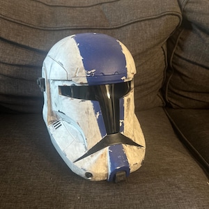 May include: A white and blue helmet with black visor and accents. The helmet has a weathered appearance with blue stripes and a dark visor. The helmet is likely a costume or collectible item.