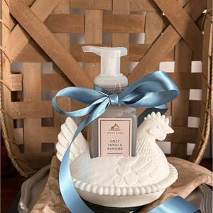 May include: A white ceramic hen-shaped soap dispenser holding a bottle of Cozy Vanilla Almond soap. The dispenser is decorated with a blue satin ribbon. The background features a woven basket.