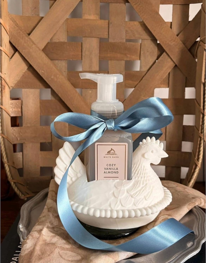 May include: A white ceramic hen-shaped soap dispenser holding a bottle of Cozy Vanilla Almond soap. The dispenser is decorated with a blue satin ribbon. The background features a woven basket.
