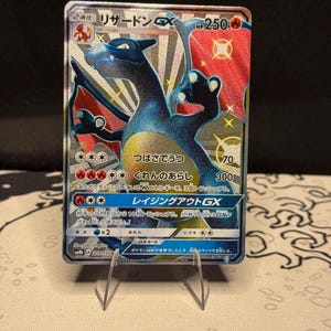 May include: A holographic Charizard GX trading card. The card features a blue and orange dragon with Japanese text. The card is displayed on a clear plastic stand against a dark background. The card has a HP of 250.