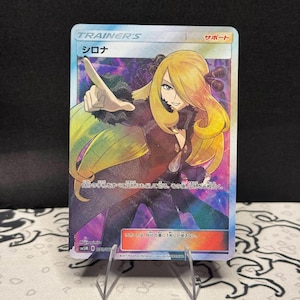 May include: A Pokémon trading card featuring the character "Sirona" with Japanese text. The card has a colorful illustration of a woman with blonde hair and a black outfit. The card is held by a clear plastic stand.