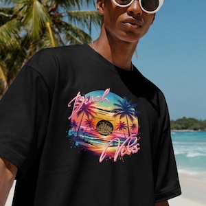 Puede incluir: Camiseta negra con un gr&aacute;fico colorido que dice "Beach Vibes". El dise&ntilde;o presenta una puesta de sol, palmeras y una paleta de colores vibrantes. La camiseta se usa en una playa con palmeras y agua azul en el fondo.
