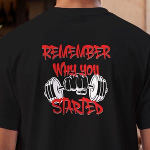 Könnte beinhalten: Ein schwarzes Kurzarm-T-Shirt mit einem Grafikdruck auf der Rückseite. Das Design zeigt eine weiße Illustration einer Faust, die eine Hantel hält, umgeben von dem roten Schriftzug "REMEMBER WHY YOU STARTED" in einer fetten Graffiti-Schrift.