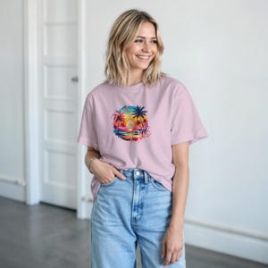 Puede incluir: Camiseta rosa claro con un dise&ntilde;o gr&aacute;fico circular de una puesta de sol, palmeras y la palabra "Paradise". El dise&ntilde;o es colorido, con tonos de azul, naranja y rosa.
