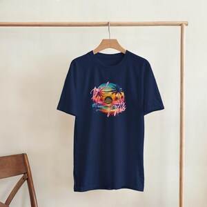 Puede incluir: Camiseta azul marino con un dise&ntilde;o de puesta de sol colorido, palmeras y el texto "Good Vibes". El gr&aacute;fico circular presenta colores brillantes como el rosa, naranja y azul.