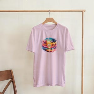 Puede incluir: Camiseta rosa claro con un gr&aacute;fico colorido de una puesta de sol sobre el oc&eacute;ano, palmeras y la palabra "Aloha". El dise&ntilde;o presenta tonos de naranja, rosa, azul y amarillo. La camiseta est&aacute; colgada en una percha de madera.
