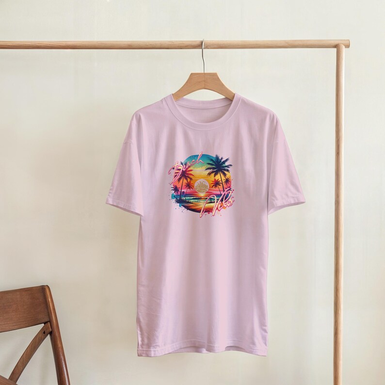 Puede incluir: Camiseta rosa claro con un gr&aacute;fico colorido de una puesta de sol sobre el oc&eacute;ano, palmeras y la palabra "Aloha". El dise&ntilde;o presenta tonos de naranja, rosa, azul y amarillo. La camiseta est&aacute; colgada en una percha de madera.