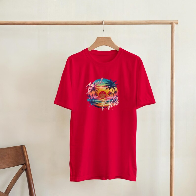 Puede incluir: Camiseta roja con un dise&ntilde;o gr&aacute;fico circular de una puesta de sol sobre una playa con palmeras. El dise&ntilde;o incluye el texto "Beach Vibes". La camiseta est&aacute; colgada en una percha de madera.