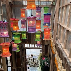 Könnte beinhalten: Eine Ansammlung farbenfroher, zylindrischer Pendelleuchten mit aufwendigen Webmustern. Die Lampen in Orange-, Rosa-, Lila-, Grün- und Blautönen sind an einer Holzdecke aufgehängt und beleuchten einen rustikalen Innenraum. Die Lampenschirme haben einen ausgestellten Sockel.