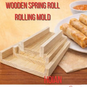 Puede incluir: Molde de madera para enrollar rollitos de primavera. El molde de madera de color claro es rectangular con secciones elevadas. El texto "Wooden Spring Roll Rolling Mold" es rojo. Un plato de rollitos de primavera y salsa está en el fondo.