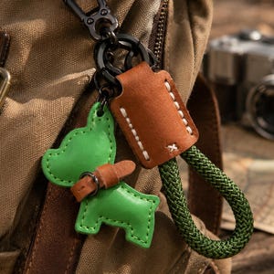 Peut inclure: Un porte-clés composé d'une breloque en cuir vert vif en forme de chien avec un petit collier en cuir marron, attaché à une boucle en cuir marron à coutures blanches et une boucle en corde vert foncé texturée, le tout relié par un mousqueton noir.