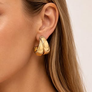 May include: Gold-toned hoop earrings with a double-layered design. The earrings have a polished, reflective surface, and a rounded shape. The earrings are shown on a person's ear, highlighting their size and style. These earrings are a classic jewelry accessory.