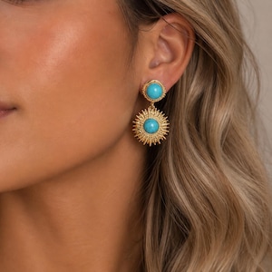 Puede incluir: Pendientes dorados con una piedra azul turquesa en el centro. El pendiente superior es un círculo pequeño, mientras que el pendiente inferior tiene un diseño de sol. Los pendientes cuelgan de la oreja.