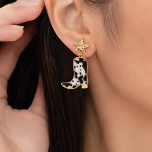 May include: Gold-tone star and cowboy boot earrings. The star-shaped stud is connected to a dangling boot with a black and white cowhide pattern. The boot has a gold-colored heel and trim. The earrings are worn on a person's ear.