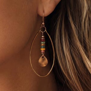 May include: A copper-toned dangle earring featuring a large oval hoop frame. Suspended from the top of the hoop is a vertical stack of small, multi-colored cylindrical beads in shades of red, teal, yellow, and copper, ending in a small teardrop charm.