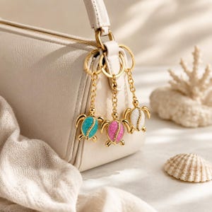 Könnte beinhalten: Drei goldfarbene Schildkröten-Schlüsselanhänger mit türkisfarbenen, rosa und weißen Emaille-Schalen hängen an einer weißen Handtasche. Die Schlüsselanhänger sind an goldenen Ringen befestigt. Eine Muschel und Korallen befinden sich im Hintergrund.