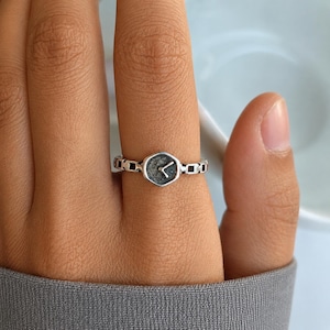 May include: A silver ring with a watch face design. The ring's band has a chain-like pattern, and the watch face is a hexagon shape with a dark grey surface and watch hands. The ring is worn on a finger.