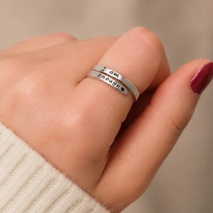 Peut inclure: Une bague argentée avec l'inscription "I am enough". La bague est ornée d'un petit cœur après le mot "enough". La bague est portée au doigt.