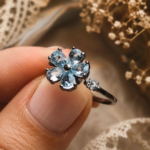 May include: A dark metal ring featuring a flower-shaped centerpiece composed of five pear-cut light blue gemstones. A single small, round, clear gemstone is set into the thin band adjacent to the floral arrangement, creating a delicate design.