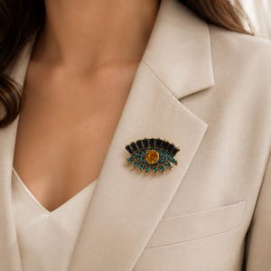 May include: An eye-shaped brooch featuring a central amber-colored circular stone, surrounded by small teal crystals and a row of black rectangular stones along the top edge, all set in a gold-toned metal frame pinned to a beige blazer.