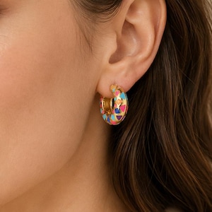 May include: Gold-tone hoop earrings with a colorful geometric pattern. The earrings feature a mosaic design with pink, blue, orange, and purple shapes. The earrings are worn on a person's ear.