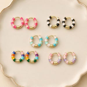 May include: A collection of colorful hoop earrings. The earrings feature gold-colored metal with enamel in various colors, including pink, black, blue, green, and purple. The earrings are arranged on a white, scalloped plate.