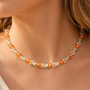 Könnte beinhalten: Eine Perlenkette mit einem sich wiederholenden Muster aus kleinen orangefarbenen und weißen blumenförmigen Perlen, durchsetzt mit winzigen durchscheinenden grünen und klaren Glasperlen, die ein durchgehendes Blumenketten-Design bilden.