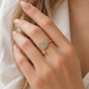Könnte beinhalten: Ein goldfarbener offener Ring mit einem kreisförmigen Motiv, das einen klaren Stein in der Mitte und einen Ring aus kleinen türkisfarbenen Steinen aufweist. Das andere Ende des offenen Rings ist mit einem kleinen, klaren, gefassten Stein versehen.