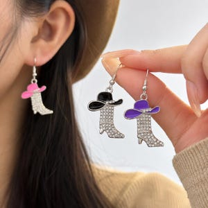 May include: Cowboy boot earrings with rhinestone detailing and colorful hat accents. The earrings feature a silver hook and are available in pink, black, and purple. A fun accessory for a country-western theme.
