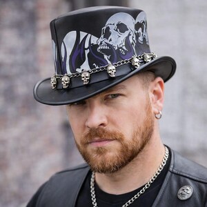 May include: Black top hat with a skull design and a silver skull chain. The hat features a black brim and a decorative skull and flame design in white and purple. A stylish accessory.