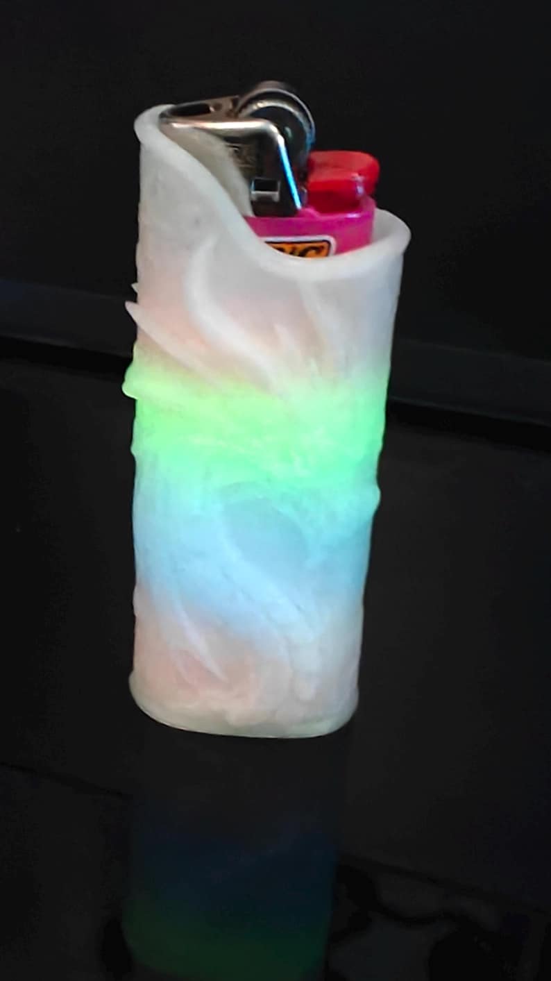 May include: A cylindrical, white, textured lighter holder with a color-changing LED light. The light transitions from pink to blue to green. A pink and black lighter is visible inside the holder.