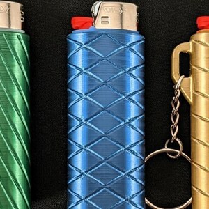 May include: Three lighters in green, blue, and gold, each with a unique textured pattern. The lighters have a red button and a silver metal top. Each lighter has a small handle and a keychain attachment.