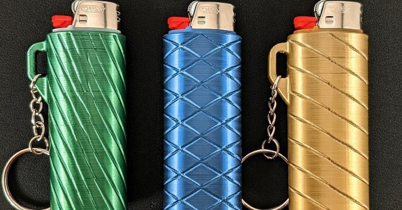 May include: Three lighters in green, blue, and gold, each with a unique textured pattern. The lighters have a red button and a silver metal top. Each lighter has a small handle and a keychain attachment.