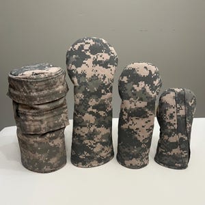 Puede incluir: Un conjunto de fundas para palos de golf y un sombrero, todos con un patrón de camuflaje digital en tonos verdes, beige y grises. Las fundas varían en tamaño y forma, diseñadas para adaptarse a diferentes tipos de palos. El sombrero es una gorra de estilo militar.