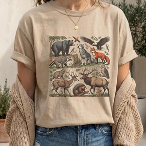 May include: Sandstone-colored t-shirt featuring a vintage-style illustration of various forest animals, including a bear, fox, and deer. The design is printed on a soft, neutral-toned tee, perfect for nature lovers.