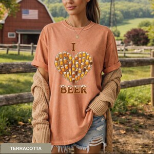 May include: Terracotta t-shirt featuring a heart made of beer glasses, with the words "I" and "BEER" above and below the heart. The shirt is worn with a beige cardigan and denim shorts. A red barn and a wooden fence are in the background.