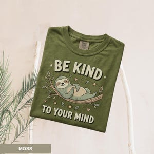 May include: Moss green t-shirt with the text "BE KIND TO YOUR MIND" and a cartoon sloth illustration. The sloth is lying on a tree branch. The shirt is a Comfort Colors brand.