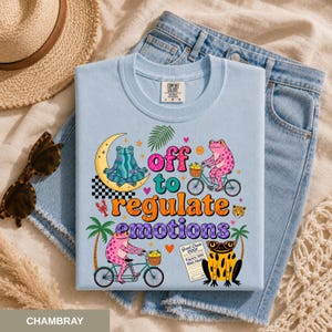 Può includere: T-shirt azzurra con una grafica colorata raffigurante rane, biciclette e la scritta "off to regulate emotions". Il design include una luna crescente, palme e un motivo a scacchiera. La maglietta è esposta su denim.