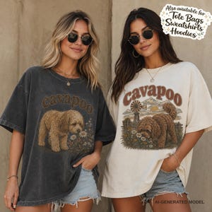 May include: Two short-sleeve t-shirts, one charcoal grey and one cream, each featuring a graphic of a brown Cavapoo dog in a field of flowers. The cream shirt includes a windmill in the background. Text on the shirts reads "cavapoo". A sticker in the corner reads "Also available for Tote Bags Sweatshirts Hoodies".