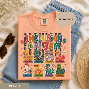 May include: A peach-colored t-shirt featuring the text INCLUSION IS MY JAM in colorful letters. Below the text are eight illustrated ducks, each with a unique pattern and a label: diversity, acceptance, educate, compassion, advocate, connection, ability, and empathy.