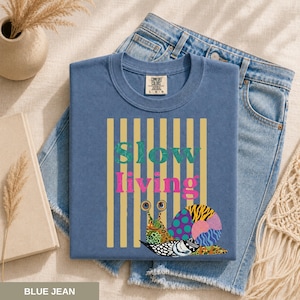 Può includere: Una maglietta blu con le parole "slow living" in verde e rosa e una grafica di lumaca colorata. La maglietta è piegata su un paio di jeans blu. La maglietta ha un'etichetta Comfort Colors.